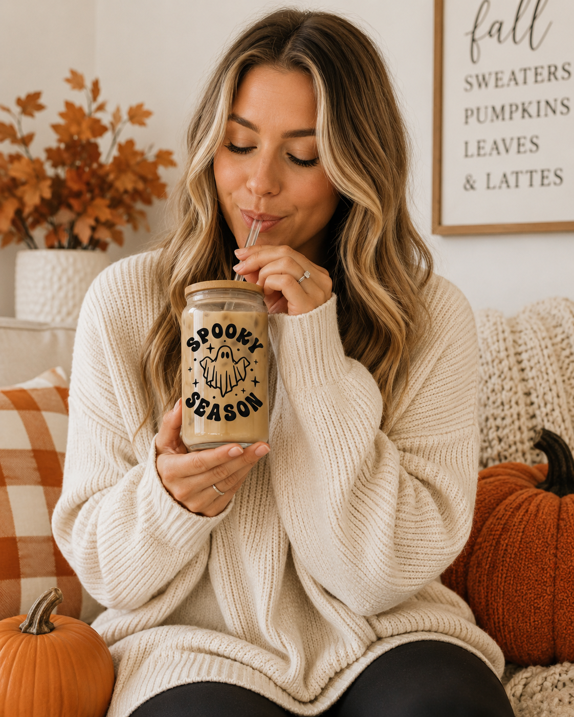 A woman in a cozy cream knit sweater holds a clear glass mason jar with a straw, featuring a black graphic that reads 'SPOOKY SEASON' with a pumpkin and ghost illustration. She is seated on a couch with pumpkins and fall decor in the background.