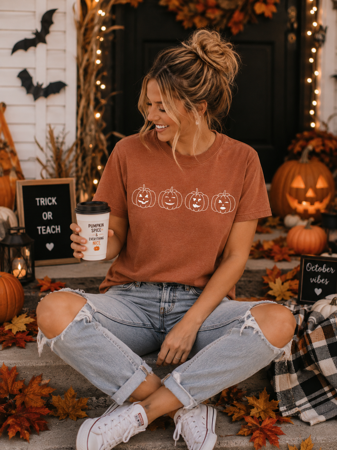 A woman sitting on steps in a Halloween-themed outdoor setting, wearing a rust-orange crewneck t-shirt with a white pumpkin graphic, ripped light-wash jeans, and white sneakers, holding a coffee cup.