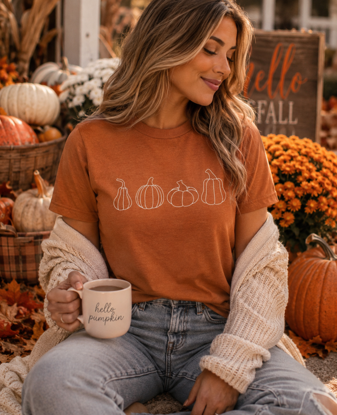 A woman wearing a rust-orange graphic t-shirt with white pumpkin line drawings, styled with a cream knit cardigan and light-wash jeans. She is holding a white ceramic mug that says 'hello pumpkin' and sitting in a cozy autumn-themed setting with pumpkins, fall leaves, and a 'hello fall' sign.